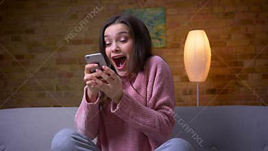 Closeup shoot of young pretty brunette caucasian female using the phone and smiling with joy sitting on the couch in a