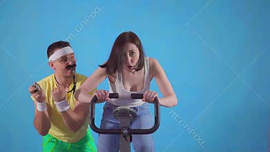 Funny man coach of 80`s with mustache trains a young woman on exercise bike on a blue background slow mo