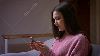 Closeup shoot of young pretty brunette caucasian female texting on the phone and laughing sitting on the couch in a cozy