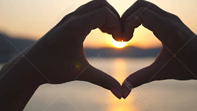Silhouette of young female hands making heart shape over sea background with beautiful sunset. Concept of summer