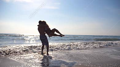 Young man hugging and spinning around his woman on sea beach. Happy boyfriend whirling girlfriend on his hands at