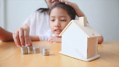 4K Mother and Asian little girl putting money coin to house piggy bank select focus on house