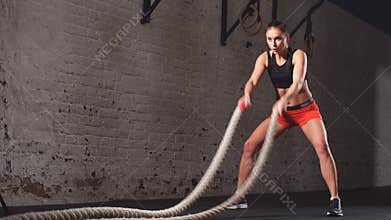 Young adult girl practising battle rope exercise during a cross fit workout at the gym,