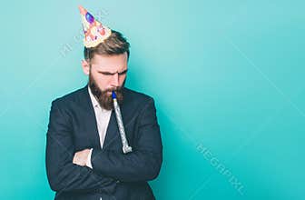 Sad guy is standing and looking down. He is upset. Man is holding a wistle in his mouth and has a birthday hat on the