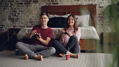 Cute young couple boyfriend and girlfriend are playing videogame holding joysticks sitting on bedroom floor at home