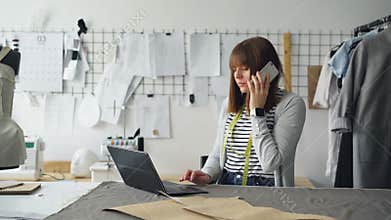 Small business owner is ordering fabric for tailor`s shop with mobile phone and working with laptop. Woman is talking