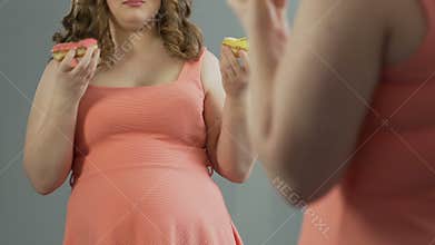 Sad fat girl crying at her reflection in mirror, eating two donuts at same time