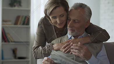 Senior man reading newspaper, woman coming from behind to hug, family morning