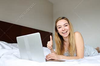 Young female person using laptop and lying on bed in morning, watching thumbs up.