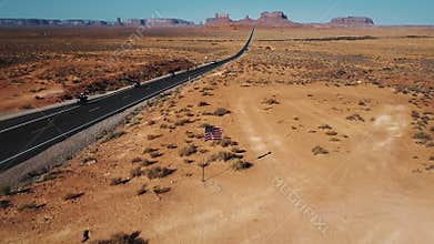Drone camera tilts up to reveal American flag waving in the middle of Monument Valley, bikers passing by on desert road.