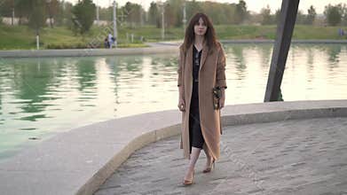 Beautiful brunette in dress and coat walking near pond in autumn park