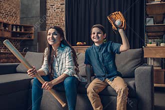 Little boy and his beautiful mother sitting on couch in living room with baseball bat and mitt while