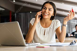 Good business talk. Cheerful young beautiful woman talking on mobile phone and using laptop with smile while sitting at