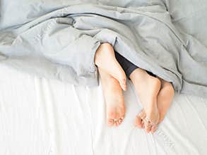 Feet of couple sleeping side by side in comfortable bed. Close up of feet in a bed under white blanket. Bare feet of a