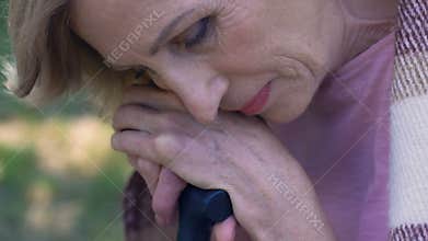 Sad elderly woman sitting in park with walking stick, feeling lonely and unhappy