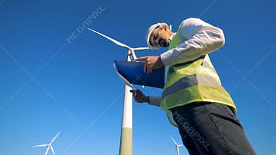Worker stands near a wind turbine, reading paper on a clipboard. Renewable electririty, green energy concept.