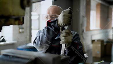 Aged male worker is adjusting machine in a workshop of factory, rotating key and using hammer