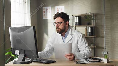 Experienced caucasian adult doctor with beard are sitting at desk and studying patients reports, against large window in