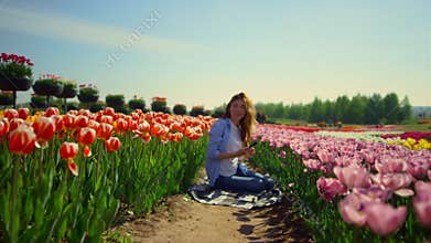 Young woman sitting in blooming flower garden with mobile phone in sunny day.