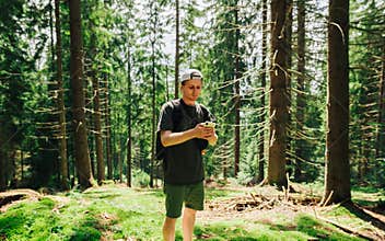 Handsome young man in casual clothes stands in the woods on a summer day with a backpack on his back and uses a smartphone with a