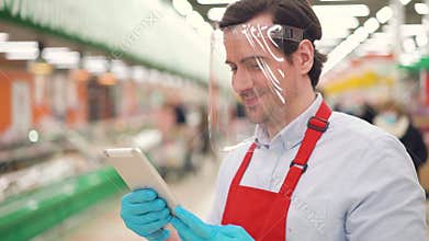 Male retail worker in plastic face shield standing in supermarket using digital tablet, taking online ecommerce orders