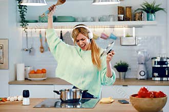 Beautiful young woman cooking while singing and listening music with headphones in the kitchen at home