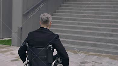 Crop view of male person in suit pushing himself on wheel chair at street. Disabled man going to work and stopping near