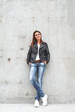 Young cute woman is standing by the wall in a leather jacket, jeans and sneakers