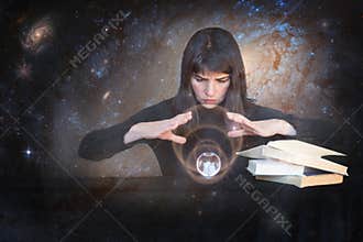 The girl holds her hands over a shining ball and looks to the future next to magic books on the background of galaxies