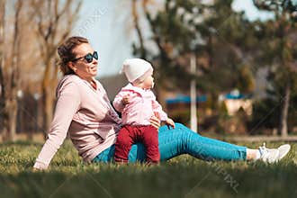 A smiling mother sits on the grass, hugging her child, enjoying the spring sun. Family outdoor recreation with children