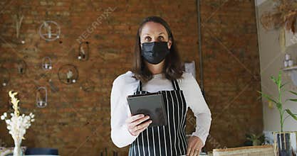 Caucasian female business owner wearing face mask and apron, using tablet, looking at camera