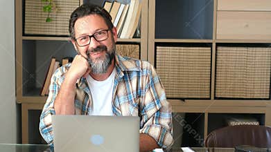 Front view handsome young businessman in eyewear working with computer remotely, sitting at wooden table in kitchen. Pleasant