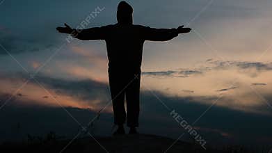 Silhouette of a happy man on top of a high mountain at sunset. A person demonstrates a sense of freedom by raising his hands up in