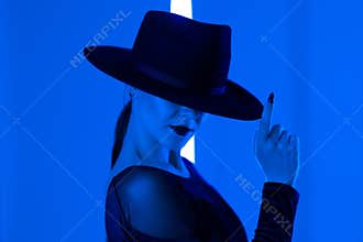 Unrecognizable sexy woman runs finger across brim of hat in dark neon studio background with colored blue light. Femme