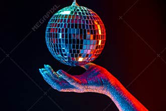 Glitter female hand holding spinning disco mirror ball under neon. Glossy silver sphere reflecting light. Retro night
