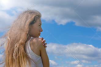 Metaphor of peace, independence and faith.Caucasian blond haired young woman hugging herself on blue summer sky background looking