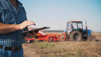 farmer is using digital tablet. close-up. on farm field, near tractor, on farm machinery backdrop. smart farming