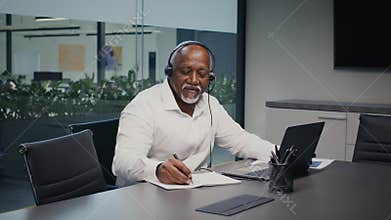 Black Businessman In Headset Taking Notes While Working With Laptop In Office