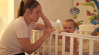 Tired sleepy mother rocking crib of her baby crying and refusing to sleep at night. Concept of parenting, parent fatigue