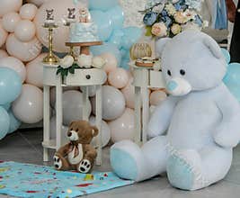 Teddy bear sits on background with different colors of balloons, birthday garland, blue and white flowers
