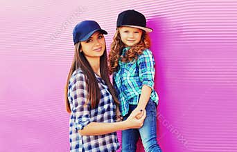Portrait of beautiful young mother with little girl child wearing checkered shirts, baseball cap in city on pink background