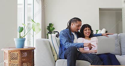 Happy biracial couple sitting on couch using laptop and talking in living room
