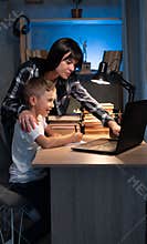 Cute boy with mother doing homework at home using laptop and books at night, online schooling