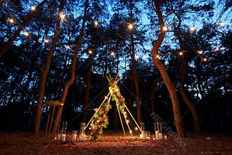 Festive string lights illumination on boho tipi arch decor on outdoor wedding ceremony venue in pine forest at night