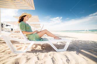 Lovely young caucasian woman freelancer in a green dress and straw hat on a schizlong under an umbrella works on her