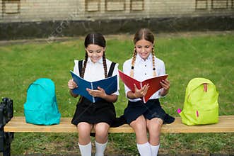 Little kids in school uniforms read library books sitting on bench outdoors, reading