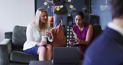 Diverse businesswomen sitting using laptop drinking coffee in office lounge
