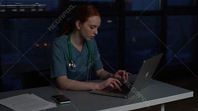 Tired depressed redhead young female doctor in blue green medical uniform working typing on laptop computer.