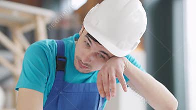 Close-up portrait of thoughtful tired young service man in hard hat wiping forehead with hand sighing. Handsome