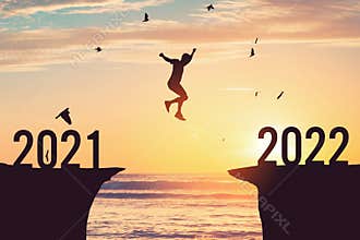 Silhouette man jumping between cliff with number 2021 to 2022 and birds flying at tropical sunset beach